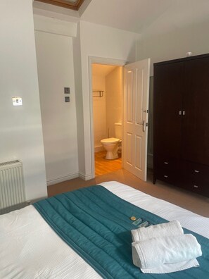 A bedroom with a bed, a dark wardrobe, a white radiator, and a bathroom visible through an open door. - Beach House - Penthouse 3 Bed Apartment (Southport)