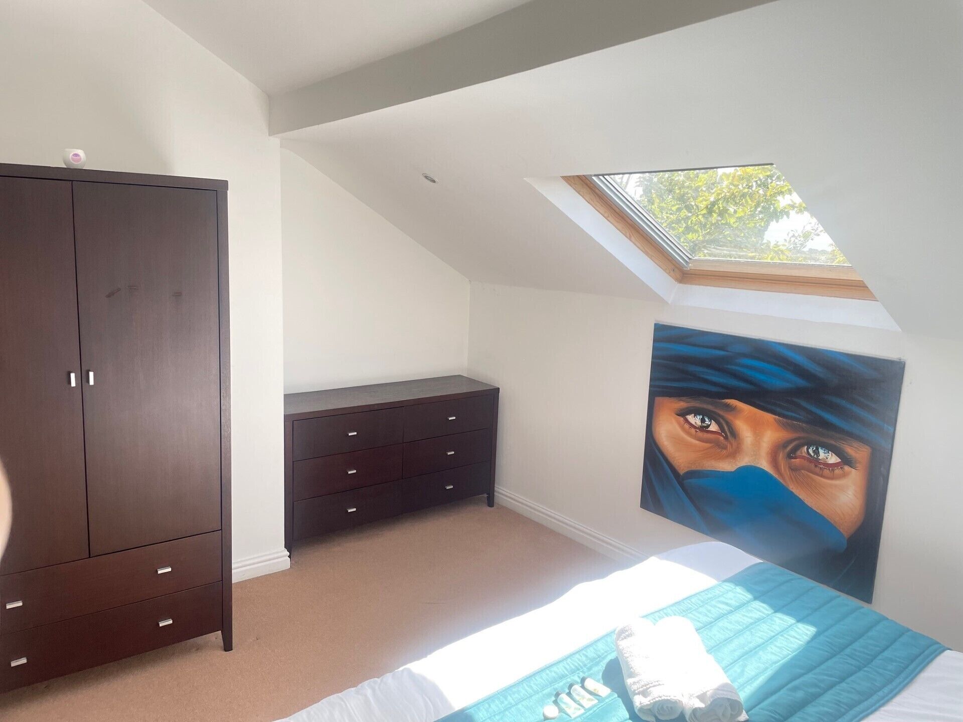 A bedroom with a sloped ceiling, a large wardrobe, a bedside table, a bed with a blue quilt, and a skylight.