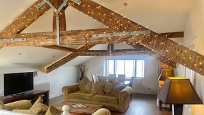 A loft-style living room with exposed wooden beams, a sofa set, a television, and a lamp. - Beach House - Penthouse 3 Bed Apartment (Southport)