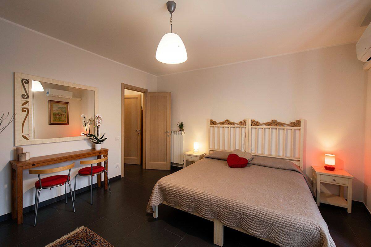 A bedroom with a bed, bedside tables, a desk with chairs, a mirror, and a door.
