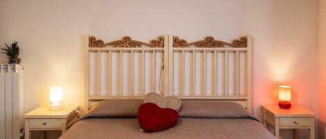 A bedroom with a bed, two bedside tables, a nightstand, a decorative headboard, and a heart-shaped pillow.