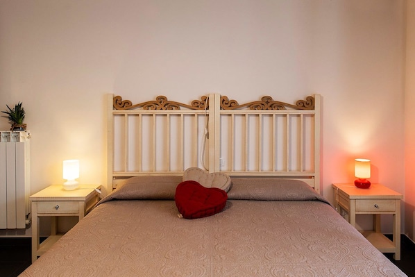 A bedroom with a bed, two bedside tables, a nightstand, a decorative headboard, and a heart-shaped pillow.