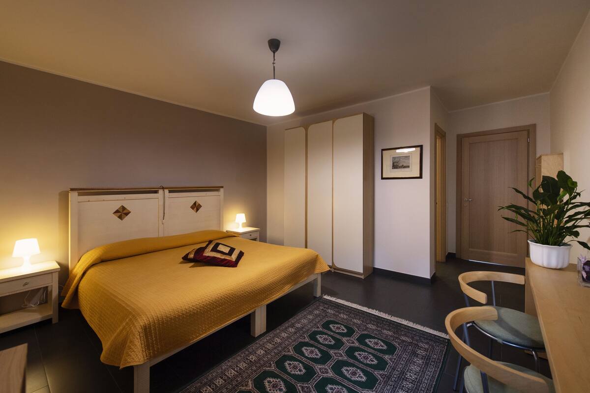 A bedroom with a bed, bedside lamps, a nightstand, a wooden chair, a potted plant, and a door.