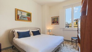 A bedroom with a bed, two pillows, a nightstand, a lamp, a chair, and two framed artworks on the wall. - Apartments Florence - Santa Maria Balcony (Firenze)