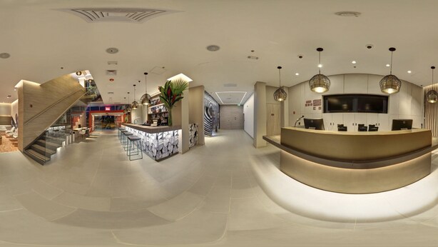 A modern hotel lobby with a curved reception desk, a staircase, and a bar area. - ibis Apartado (Apartad ó)