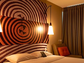 A hotel room with a large circular wall art, two wall-mounted lamps, a bed with pillows, a chair, and a curtain. - ibis Apartado (Apartadó)