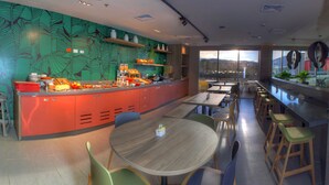 A modern restaurant with a vibrant green wall, a long counter with a variety of food items, and a seating area with round tables and chairs. - ibis Apartado (Apartadó)