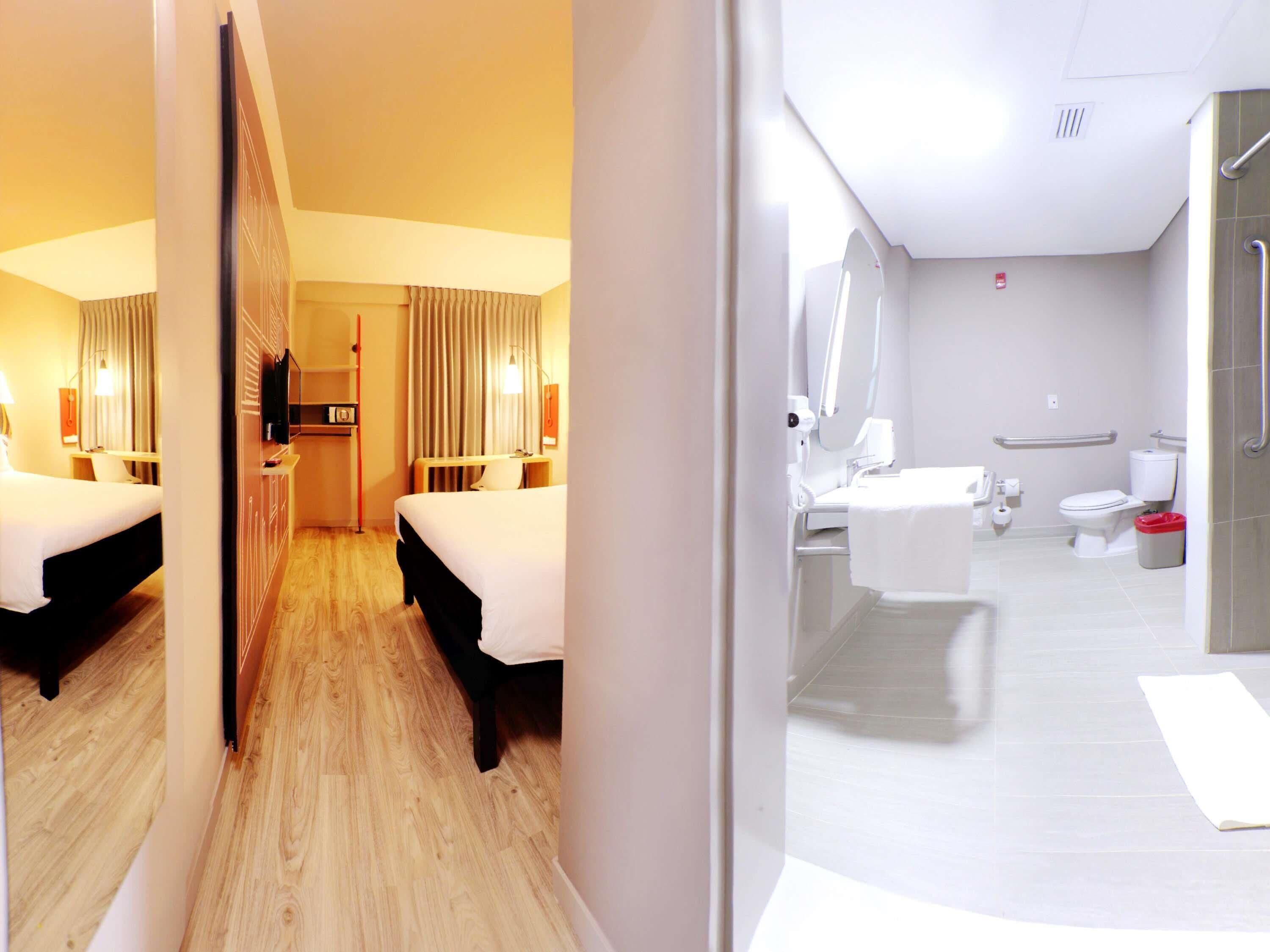 Une chambre d’hôtel avec deux lits, un bureau, un miroir et une salle de bain comprenant des toilettes, un lavabo et une douche.