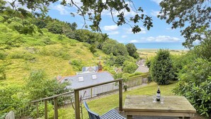 A wooden deck with metal chairs and a table, overlooking a lush green landscape and a body of water. - West Wind Holiday Home in Porlock (Porlock)