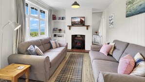 A cozy living room with two sofas, a fireplace, a bookshelf, and a large window with curtains. - West Wind Holiday Home in Porlock (Porlock)