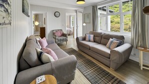 A cozy living room with two sofas, a plaid rug, and a large window with curtains. - West Wind Holiday Home in Porlock (Porlock)