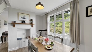 A dining area with a wooden table, chairs, a pendant light, a fireplace, and a view of greenery through large windows. - West Wind Holiday Home in Porlock (Porlock)