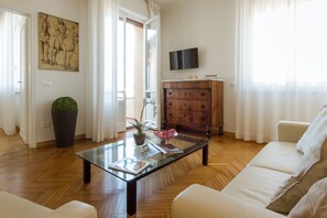 A living room with a glass coffee table, a sofa, a wooden cabinet, a television, and a painting on the wall.