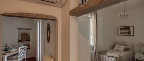 A room with a bed, a desk, and a chair. There is a radiator, a window, and a hanging light fixture.