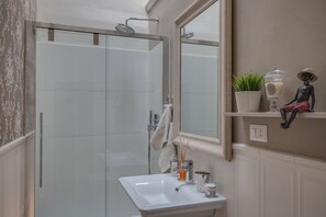 Une salle de bain avec une douche à parois de verre, un lavabo blanc, un miroir, une étagère avec des objets décoratifs et une plante en pot.