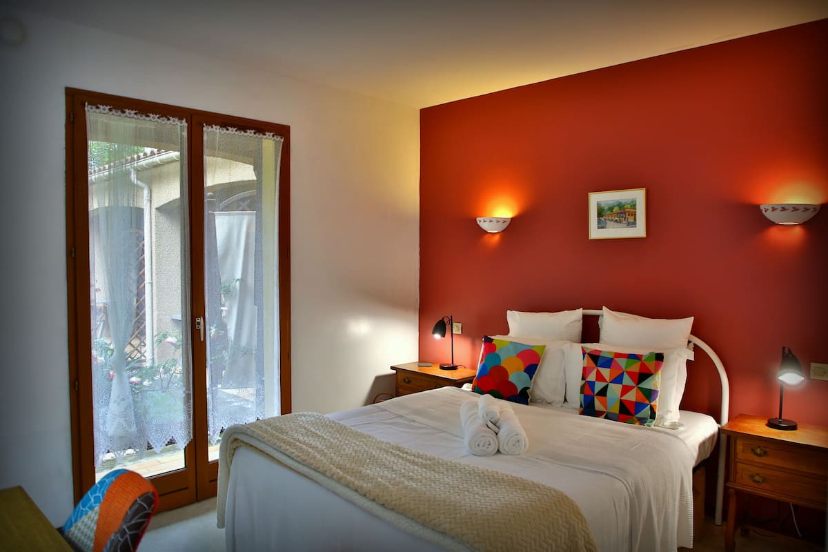A bedroom with a bed, two bedside tables, a nightstand, a window with curtains, and a red wall.
