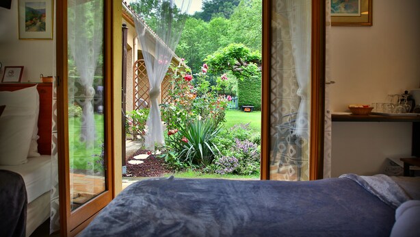 A bedroom with a large bed, a window with sheer curtains, and a view of a garden with flowers and a lawn. - Beau Vallon - Gîte et Chambres d'Hôtes (Sahorre)