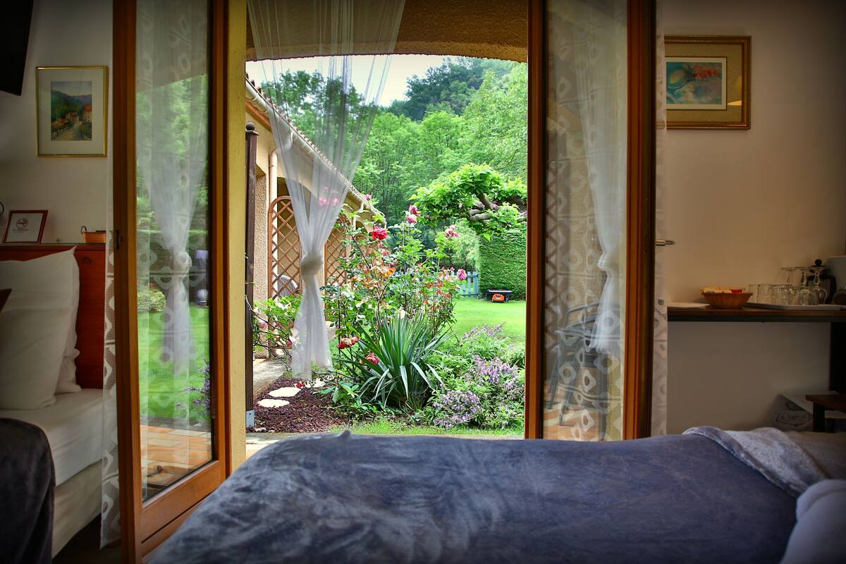 A bedroom with a large bed, a window with sheer curtains, and a view of a garden with flowers and a lawn.