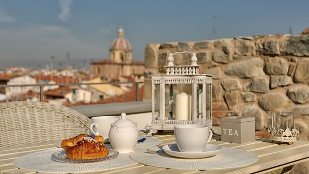 A table with a croissant, teapot, cups, and a tea box on a wicker chair with a city view in the background. - Apartments Florence - Beautiful Terrace Serragli (Firenze)