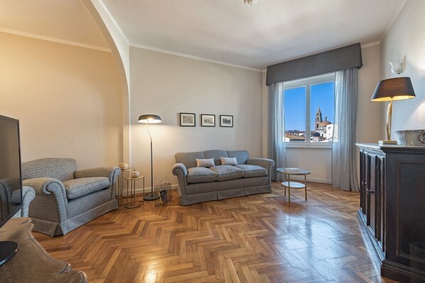 A living room with a sofa, a coffee table, a lamp, and a view of a cityscape through a window. - Apartments Florence - Carolina (Firenze)