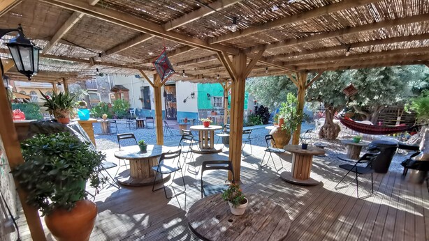 An outdoor patio area with wooden tables and chairs, potted plants, and a thatched roof structure.