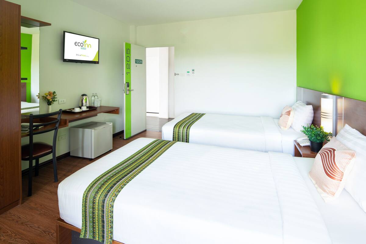 A hotel room with two beds, a desk with a chair, a small refrigerator, and a television on the wall.