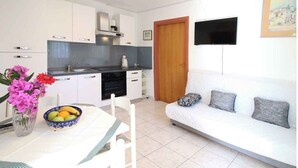 Une cuisine moderne avec des meubles blancs, une table à manger sur laquelle se trouve un saladier rempli de fruits, et un canapé blanc agrémenté de coussins à motifs.