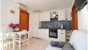 Une cuisine compacte avec des meubles blancs, un four encastré et une machine à café. On y trouve un coin repas avec une table et des chaises, ainsi qu'un lit agrémenté d'un coussin à motifs.