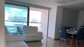 A modern living room with a white sofa, a dining area with blue chairs, and large windows offering a view of buildings. - Apartamento de Lujo Frente al Mar (Cartagena)