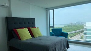 A modern bedroom with a large bed, a blue armchair, and a view of a highway and buildings. - Apartamento de Lujo Frente al Mar (Cartagena)