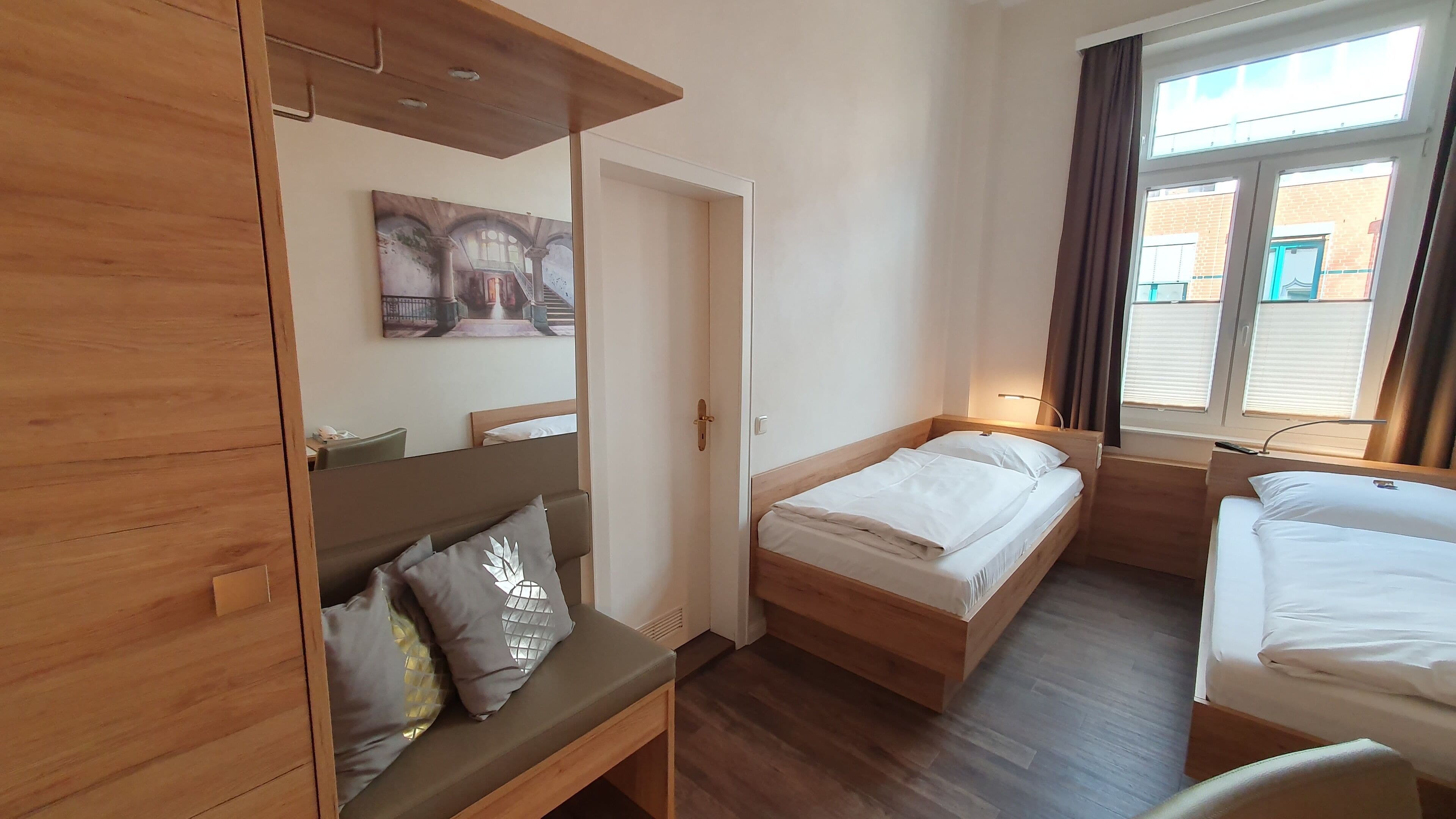 A hotel room with a bunk bed, a single bed, wooden furniture, a painting on the wall, and a window with curtains.