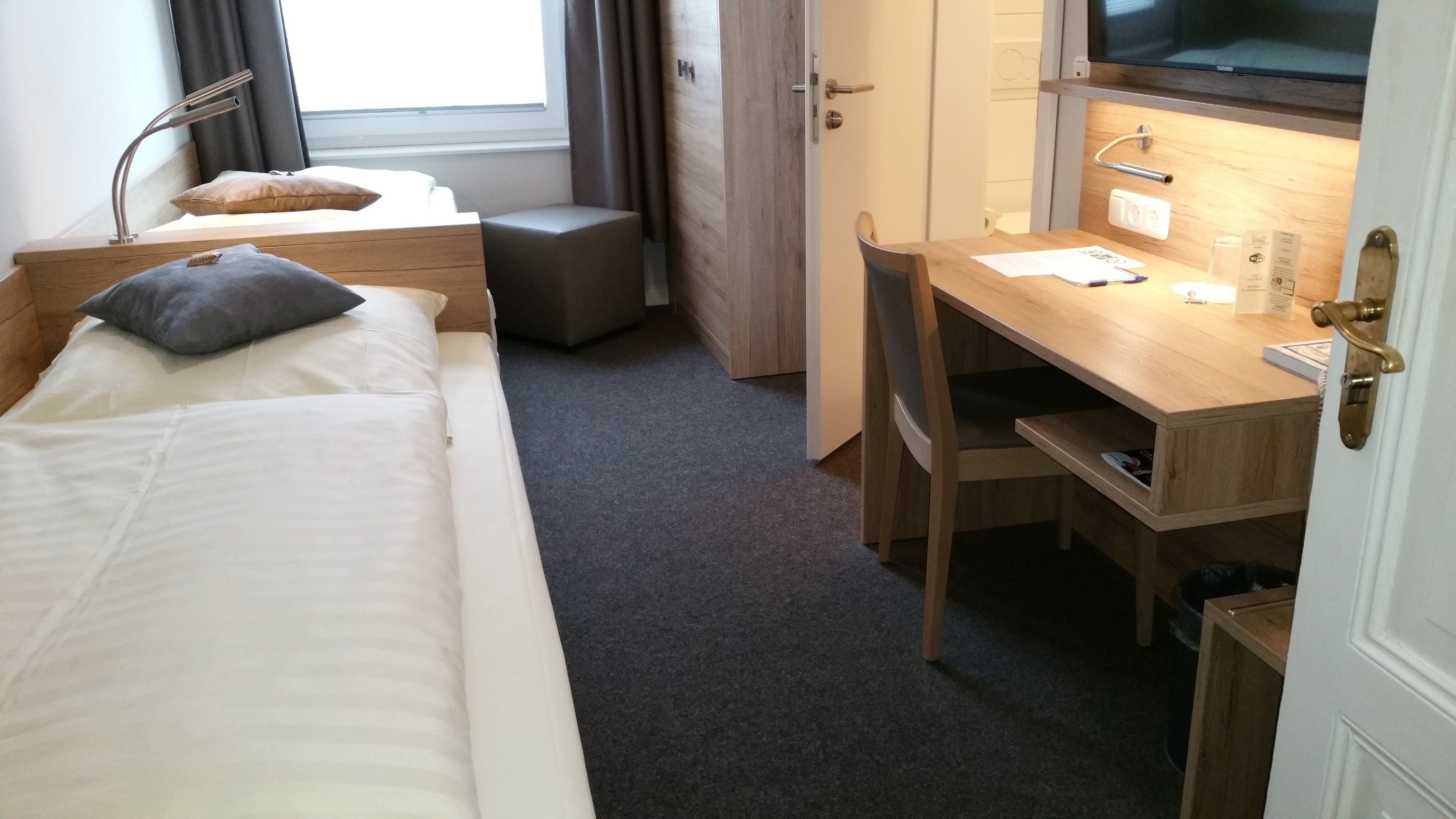A hotel room with a bed, a desk, and a chair. There is a window with curtains, a TV, and a small trash bin.