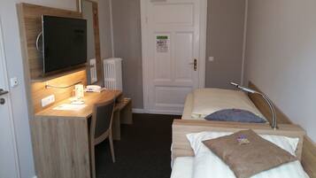 Habitación de hotel con un escritorio de madera, una cama con almohada, una mesita de noche, un televisor montado en la pared y una puerta con un cartel.