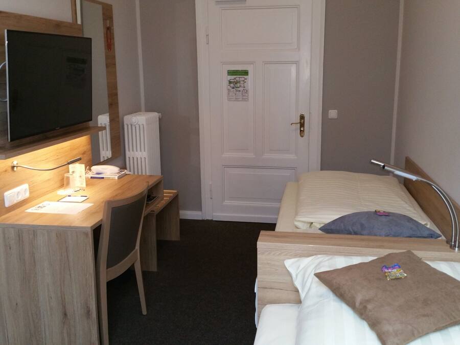 Habitación de hotel con un escritorio de madera, una cama con almohada, una mesita de noche, un televisor montado en la pared y una puerta con un cartel.