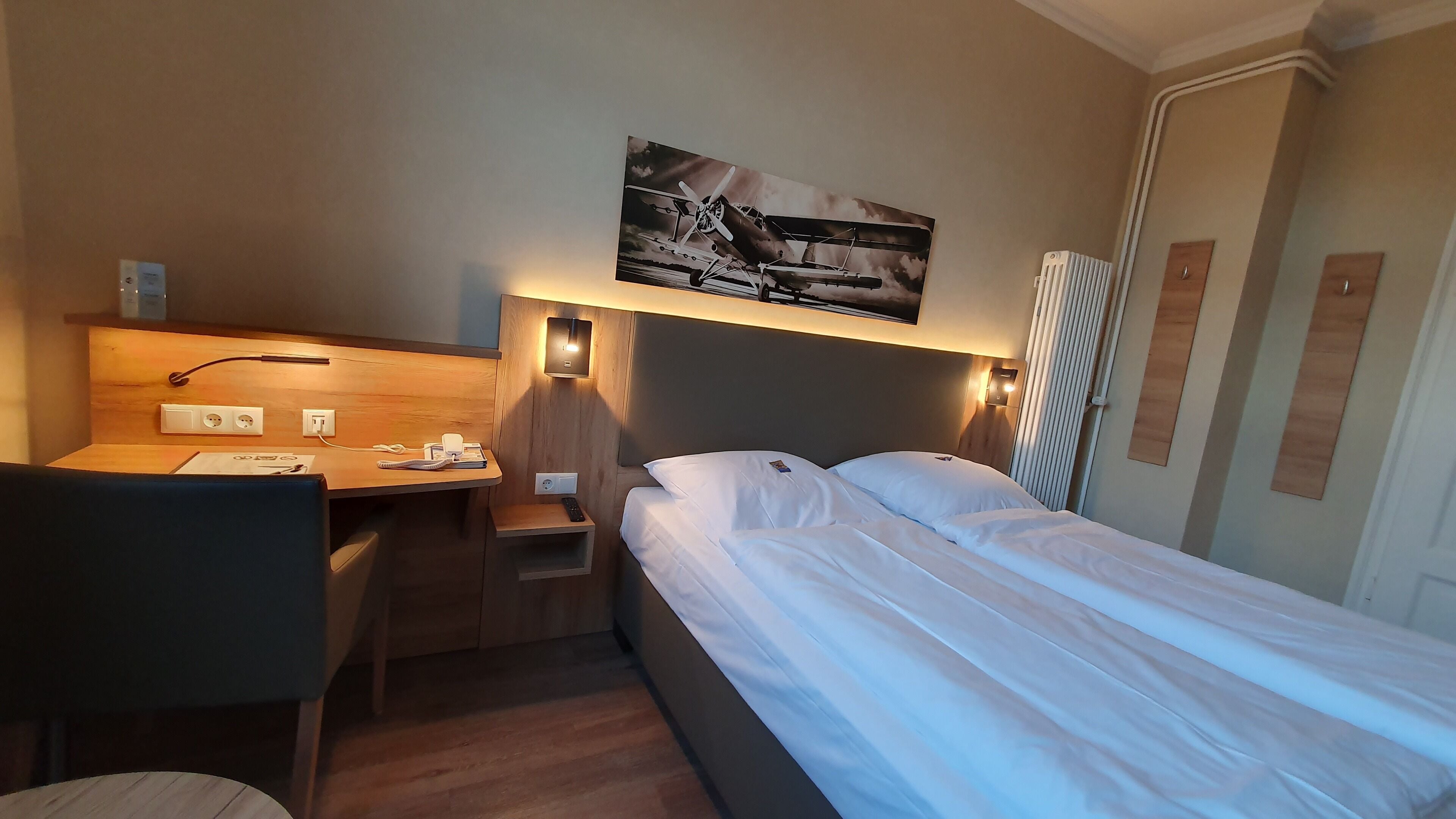 A hotel room with a bed, a desk, and a chair. There is a painting on the wall above the bed.