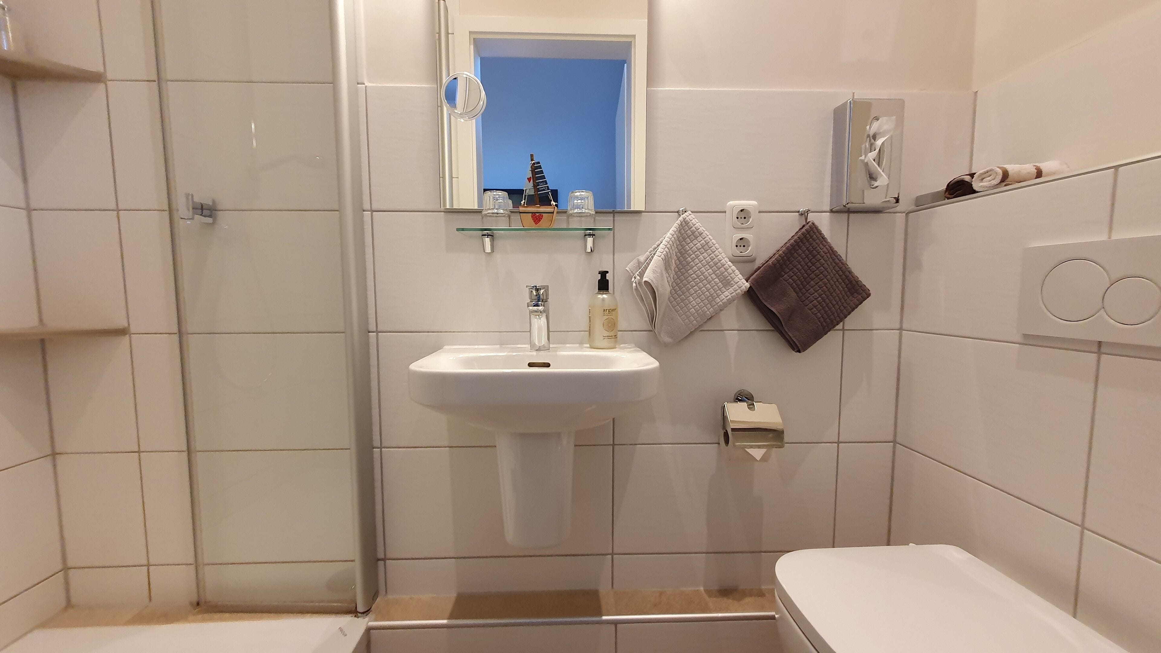 Une salle de bain avec un lavabo blanc, un miroir, une petite étagère contenant des articles de toilette, un porte-serviettes avec deux serviettes et des toilettes.