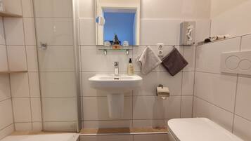 Un baño con un lavamanos blanco, un espejo, un estante pequeño con artículos de tocador, un perchero con dos toallas y un inodoro.
