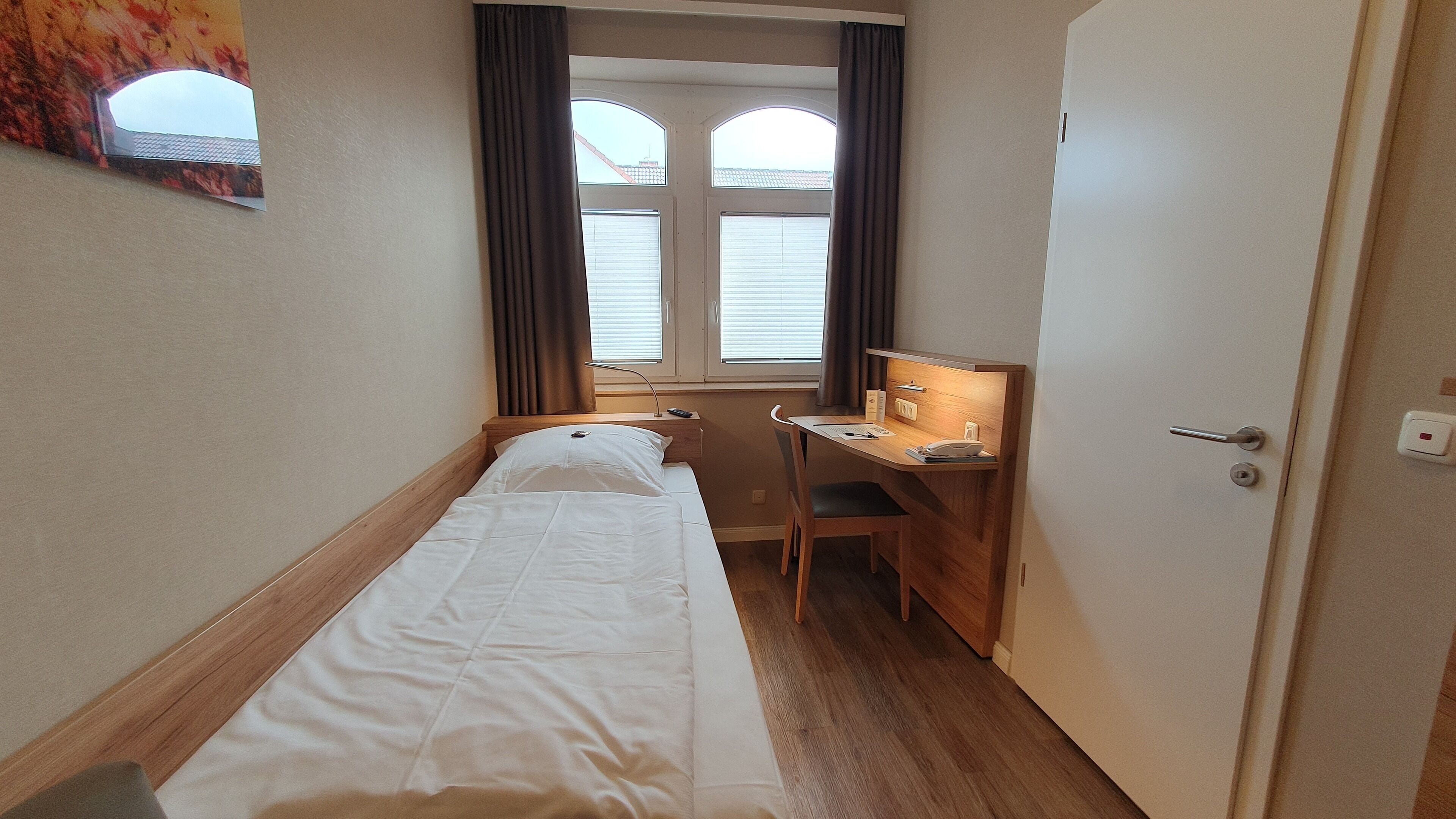 A small, single-bed room with a wooden desk and chair, a window with curtains, and a door.