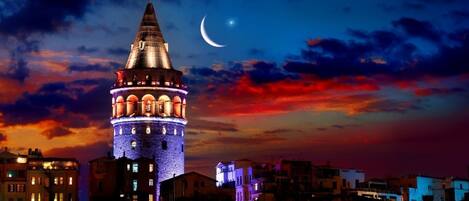 A historic tower with a conical roof, illuminated by lights, set against a twilight sky with a crescent moon.