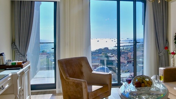 Een ruime kamer met een groot raam en uitzicht over de stad, een comfortabele fauteuil, een tafeltje met glazen blad en een decoratieve kroonluchter.