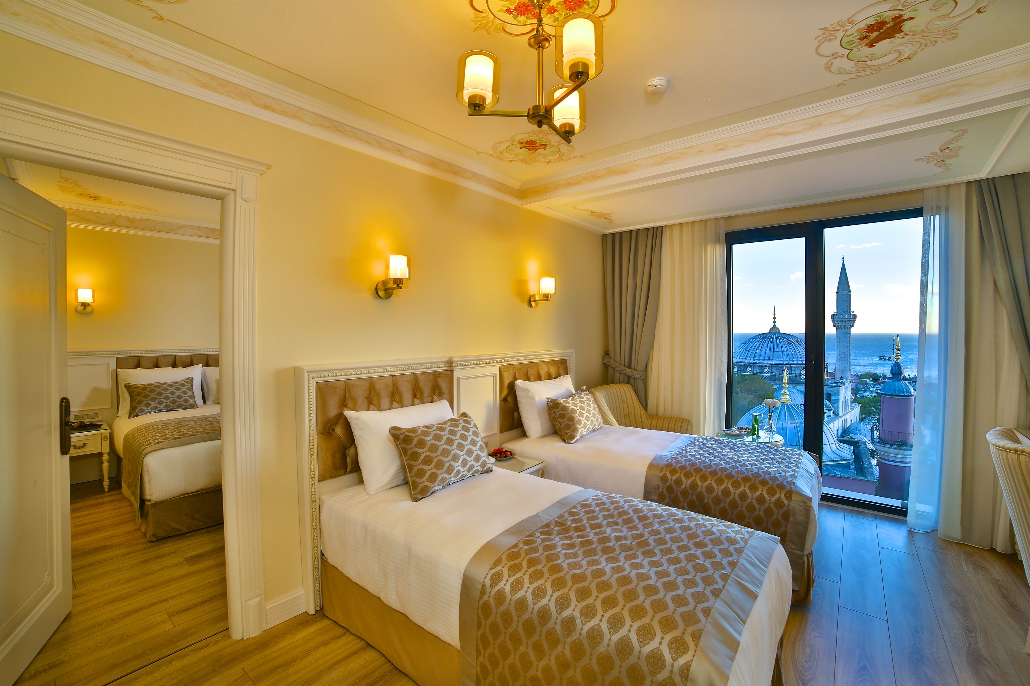 A hotel room with two beds, a chandelier, and a view of a cityscape through a large window.