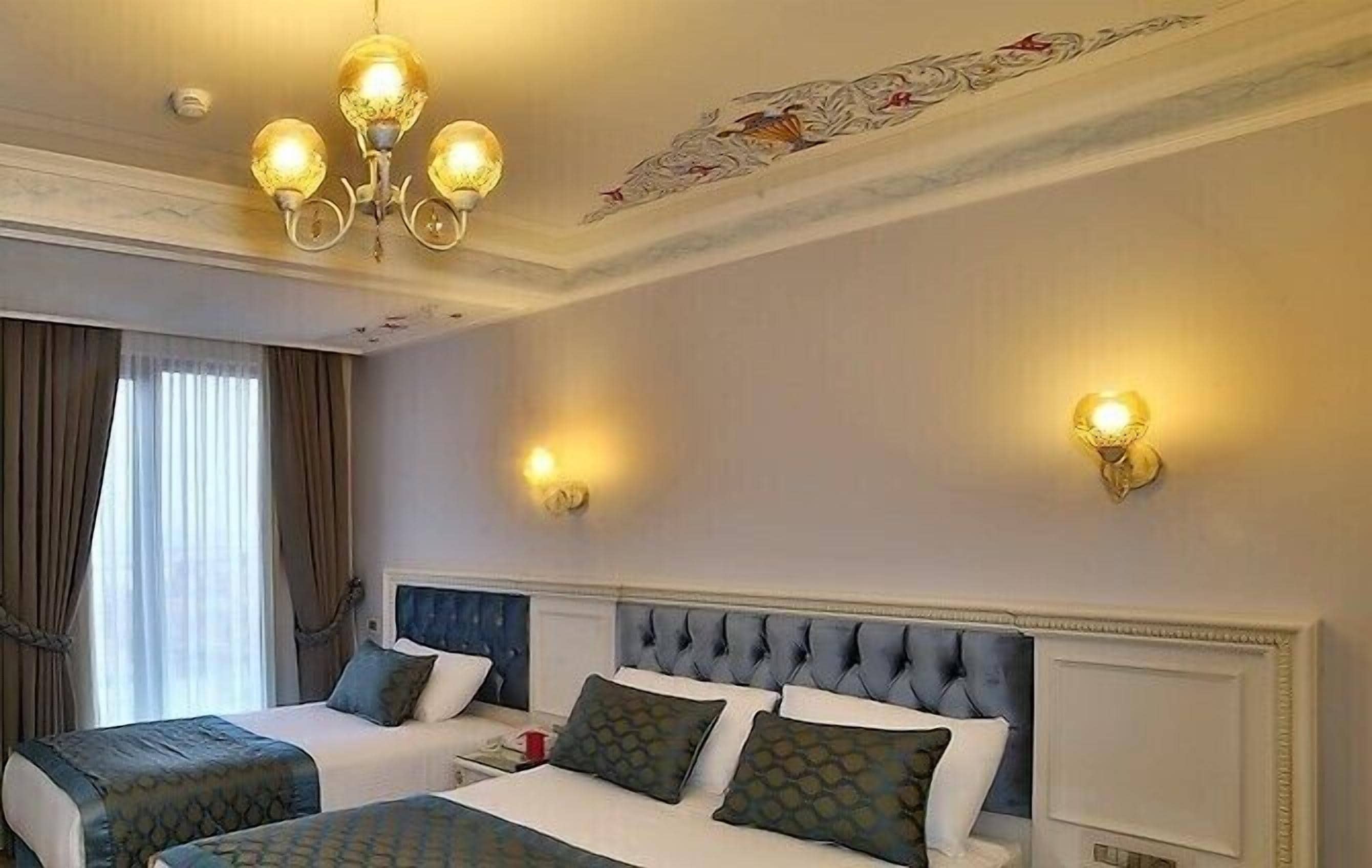 Una habitación de hotel con una cama grande, dos mesitas de noche, una araña y apliques decorativos en la pared.