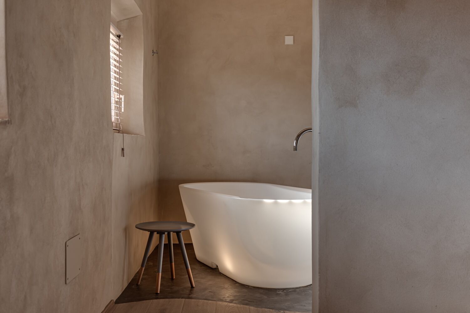 Un baño minimalista con una bañera independiente, un taburete pequeño y una ventana con persianas.