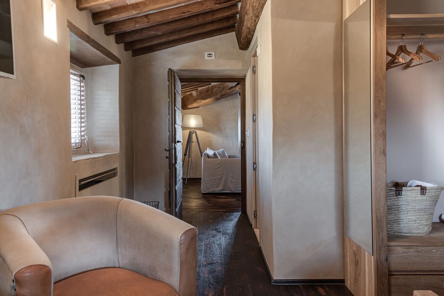 A narrow hallway with a leather armchair, wooden beams, a bed with a lamp, and a wicker basket.