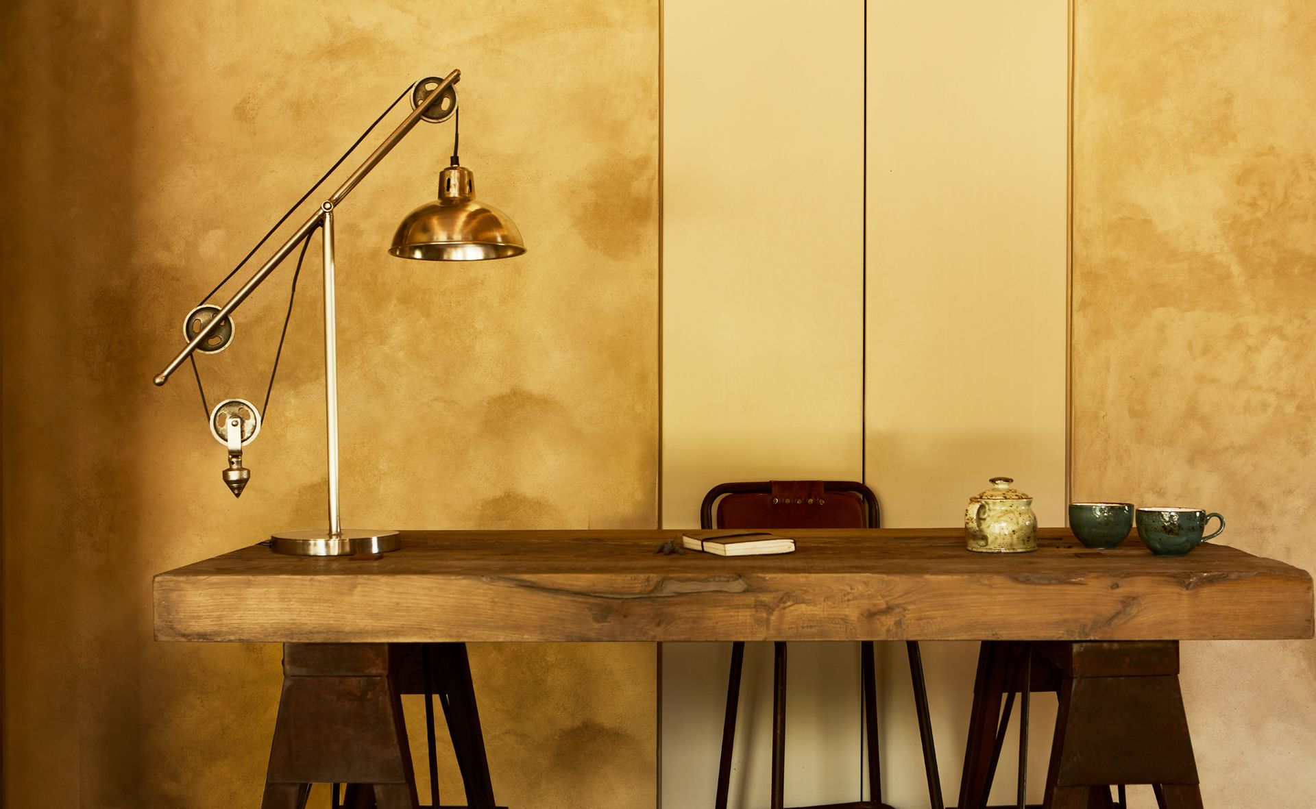 Una mesa de madera con una lámpara de escritorio de estilo vintage, una silla roja y una jarra de cerámica sobre la mesa.
