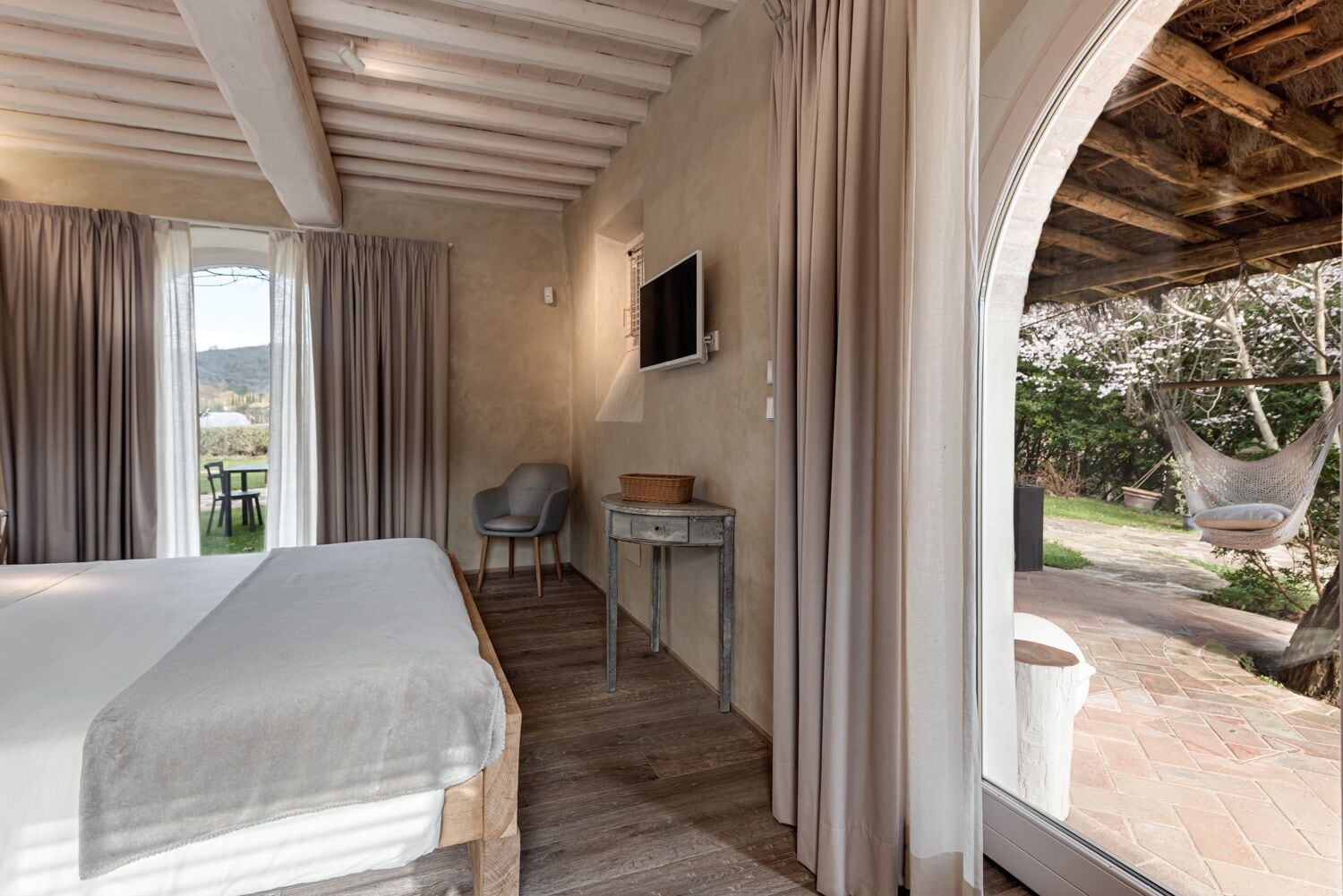 Un dormitorio con cama, una silla, una mesita y un televisor. Se ve una ventana con cortinas y vistas al exterior.