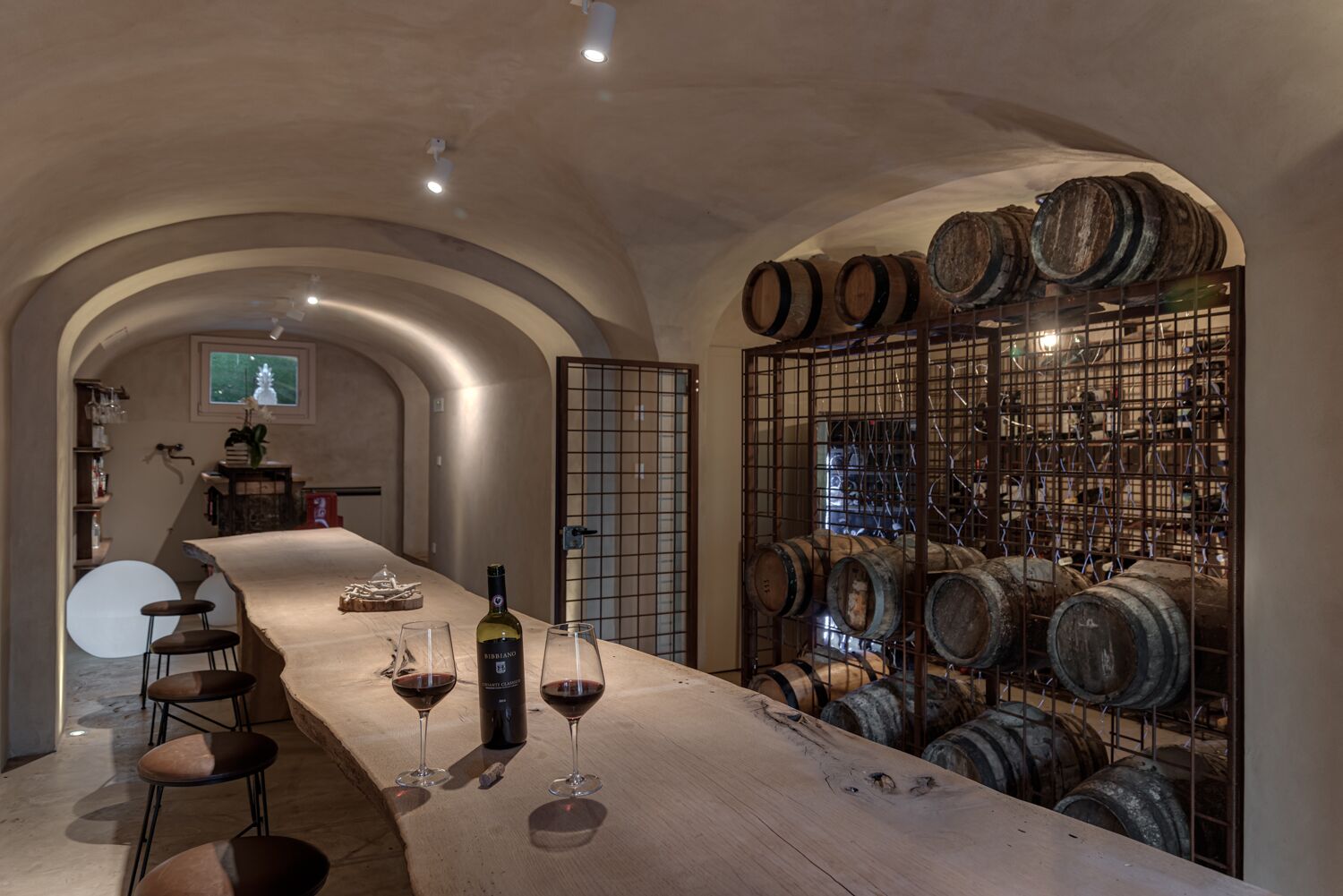 Una bodega con una larga mesa, copas de vino y una botella. Se ven barriles de madera y un estante metálico con más barriles.