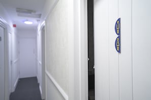 A narrow hallway with white walls and a carpeted floor. A door with a blue and yellow sign is visible on the right. - OYO Greenway Stays (Uxbridge)