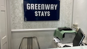 Ein Schild mit der Aufschrift "GREENWAY STAYS" in einem Raum mit einer weißen Tür, einem Metallstuhl und einem Schreibtisch mit Computer und Unterlagen.
