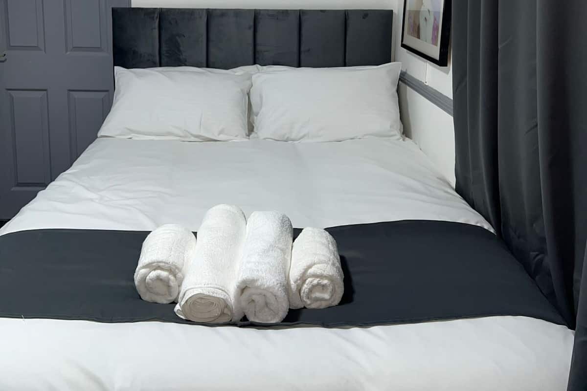 A neatly made bed with white linens and rolled towels, a dark headboard, and a grey door.